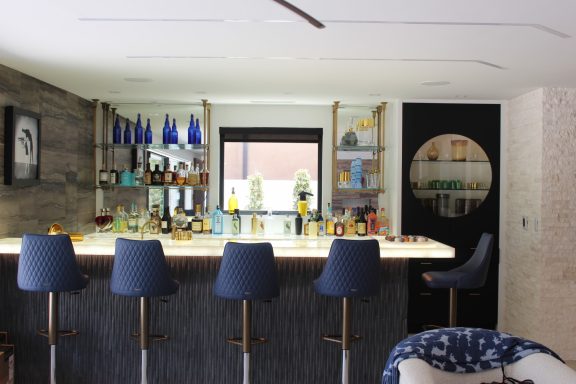 Modern home bar with blue stools, shelves of spirits, and a stylish backdrop.