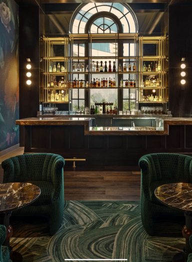 Elegant bar area with green velvet chairs, ornate decor, and a large arched window.