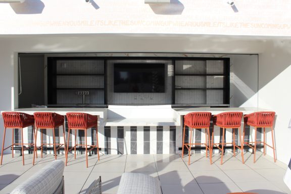 Modern outdoor bar area with high stools and a large screen above a sleek countertop.