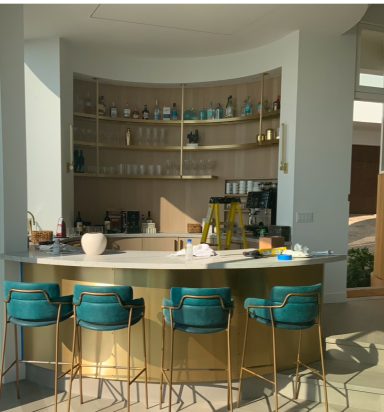 Modern bar with a gold countertop and teal chairs, surrounded by shelves of glassware.
