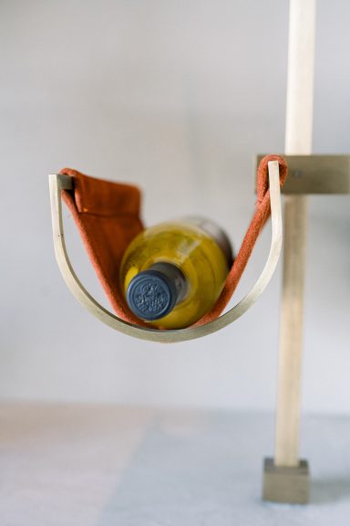 Close-up of a wine bottle cradled in soft rust-toned leather with custom brass wall-mounted supports.